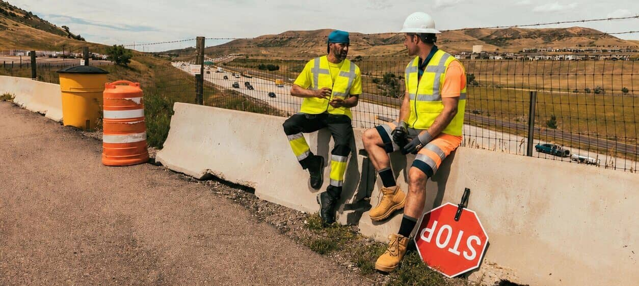 Twee arbeiders zitten op een betonnen vangrail langs de weg. Ze dragen reflecterende kleding met korte mouwen en koelvesten. Op de achtergrond is een kaal berglandschap en een bewolkte, blauwe lucht te zien.