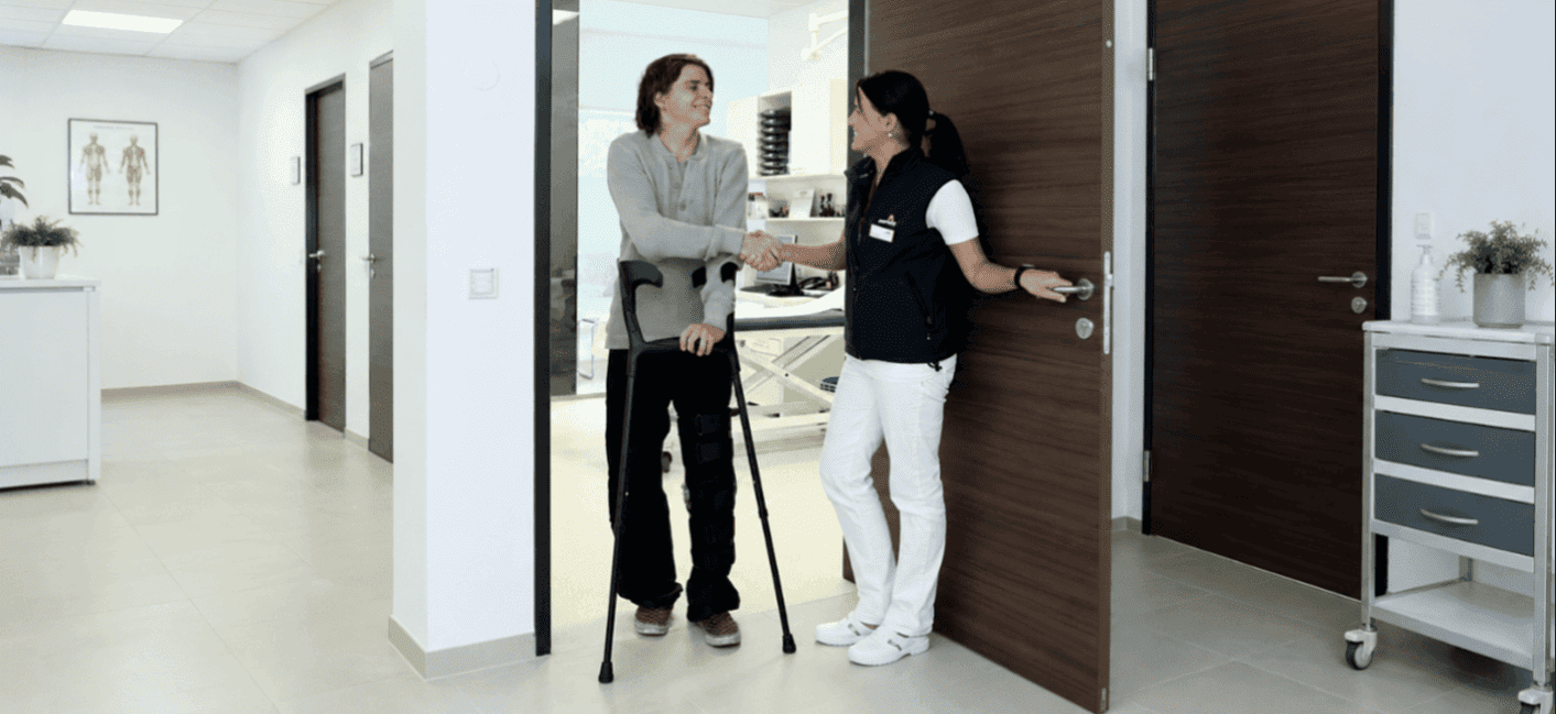Twee mensen staan in de gang van een kliniek. Een man met krukken schudt de hand van een vrouwelijke arts.