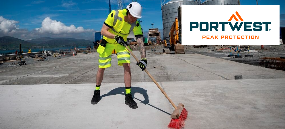 Een man in felgele werkkleding en een witte veiligheidshelm veegt de betonnen vloer voor een fabriek. Op de achtergrond zie je bergen en een blauwe lucht met kleine wolkjes. Het blauw-oranje logo van de fabrikant, Portwest, staat rechtsboven.