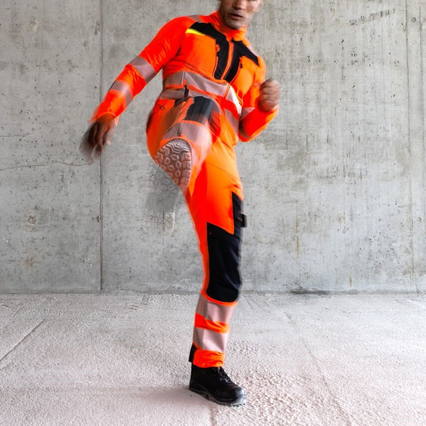 Werknemer in oranje beschermende kleding heeft één been omhoog in een trapbeweging.