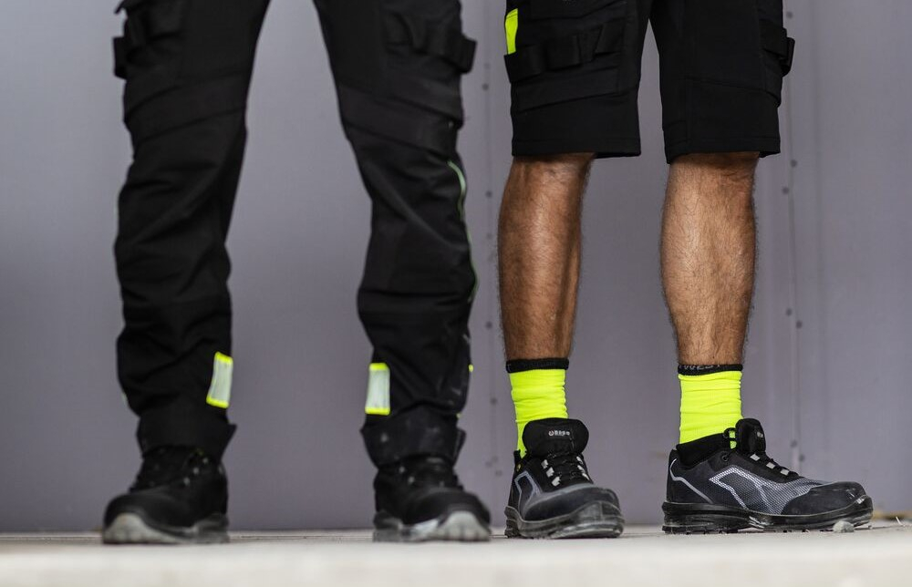 Je ziet twee paar mannenbenen: de ene man draagt een zwarte lange werkbroek, de andere een korte signaalgele werkshort. Er is een link naar onze werkbroeken toegevoegd.