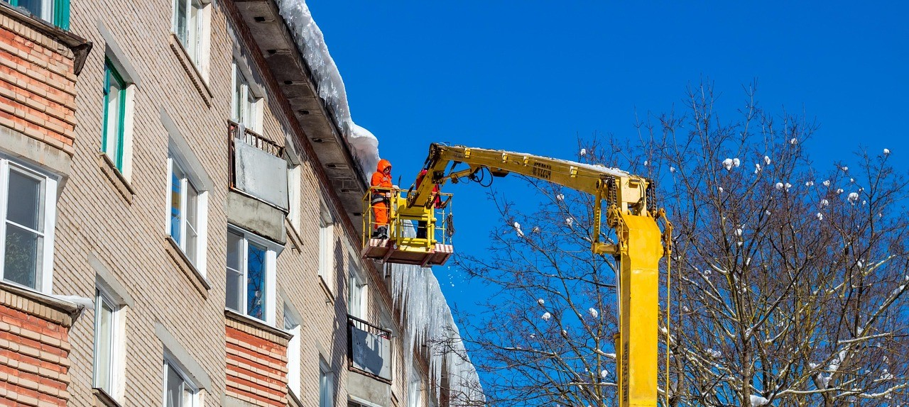 Een rij huizen met ijspegels op de nok en sneeuw op het dak. Een werker in oranje reflecterende kleding staat op een schaarhoogwerker. Rechts is een kale boom met sneeuw op de takken te zien.