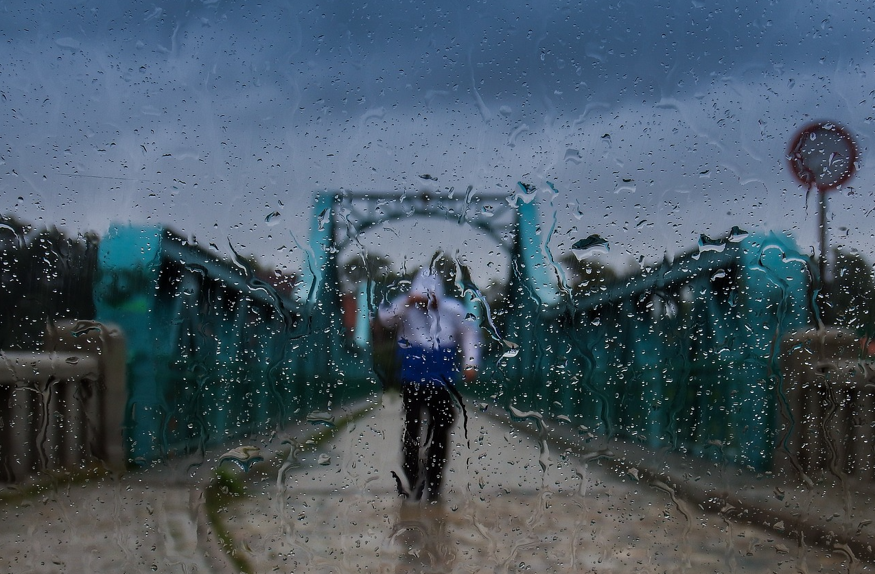 Blauwe brug tegen een grijze lucht met een persoon in regenkleding midden op de weg. Het tafereel werd gefotografeerd door een natte ruit. Er wordt een link gegeven naar onze volledige selectie praktische regenkleding.