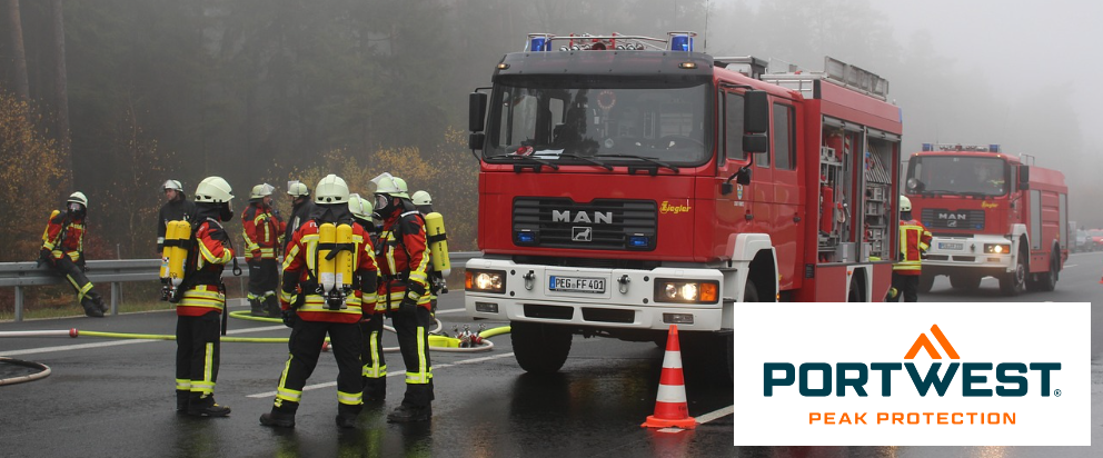 Straatbeeld met twee rode brandweerauto's en diverse brandweerlieden. Op de achtergrond zie je bomen bedekt met mist. In de rechteronderhoek staat het blauworanje Portwest-logo voor een witte rechthoek.