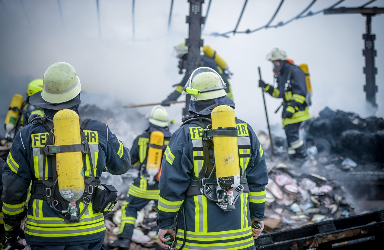 Brandweerlieden in blauwe kleding met gele waarschuwingsstrepen en helmen in een schroothoop. De bijgevoegde link leidt naar onze selectie vlamvertragende brandweerkleding.