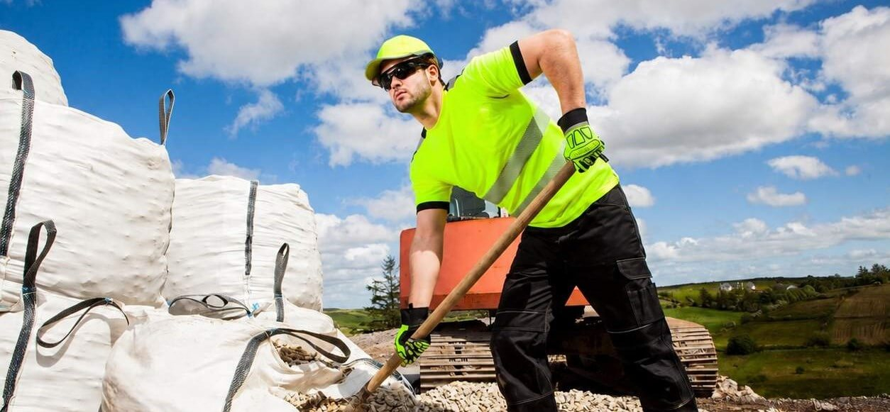 Een man in een geel shirt en een zwarte broek. Hij draagt een gele pet en een zonnebril en schept grind uit grote zakken. Op de achtergrond is een licht bewolkte blauwe lucht, een bouwplaats en groene heuvels te zien.