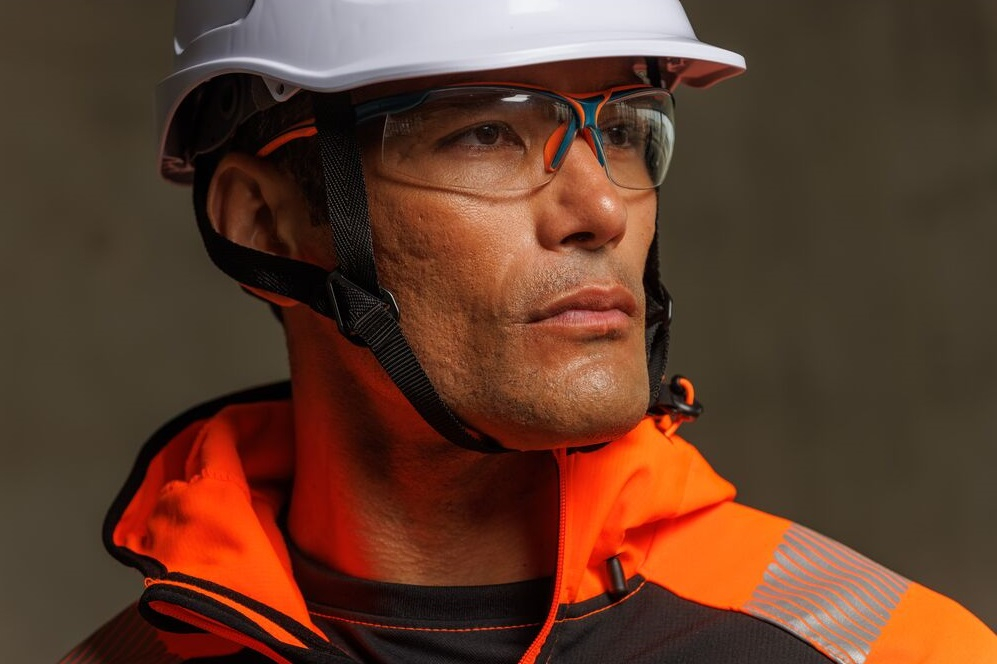 Close-up van een werknemer met een witte veiligheidshelm en heldere veiligheidsbril die geconcentreerd in de verte kijkt; draagt fel oranje-zwarte waarschuwingskleding. Een link naar onze collectie oogbescherming is toegevoegd.