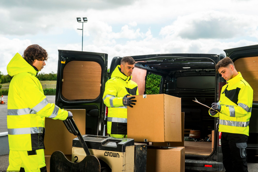 Drie mannen in felgele softshelljassen met reflecterende strepen zijn aan het werk voor een open bestelbus. Eén duwt een pallet met dozen op een palletwagen, een ander tilt een grote doos van de vrachtwagen en de derde man schrijft iets op een klembord. De scène speelt zich buiten af onder een bewolkte hemel. Een link naar onze softshelljassen is beschikbaar.