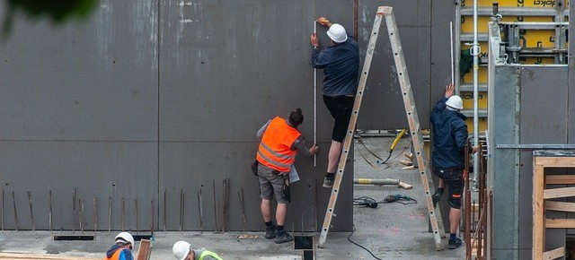 Twee werknemers zijn te zien op een bouwplaats. De ene staat op een ladder, de andere houdt hem vast. Beiden dragen een werkbroek; de ene draagt een witte helm en de andere een oranje reflecterend vest.