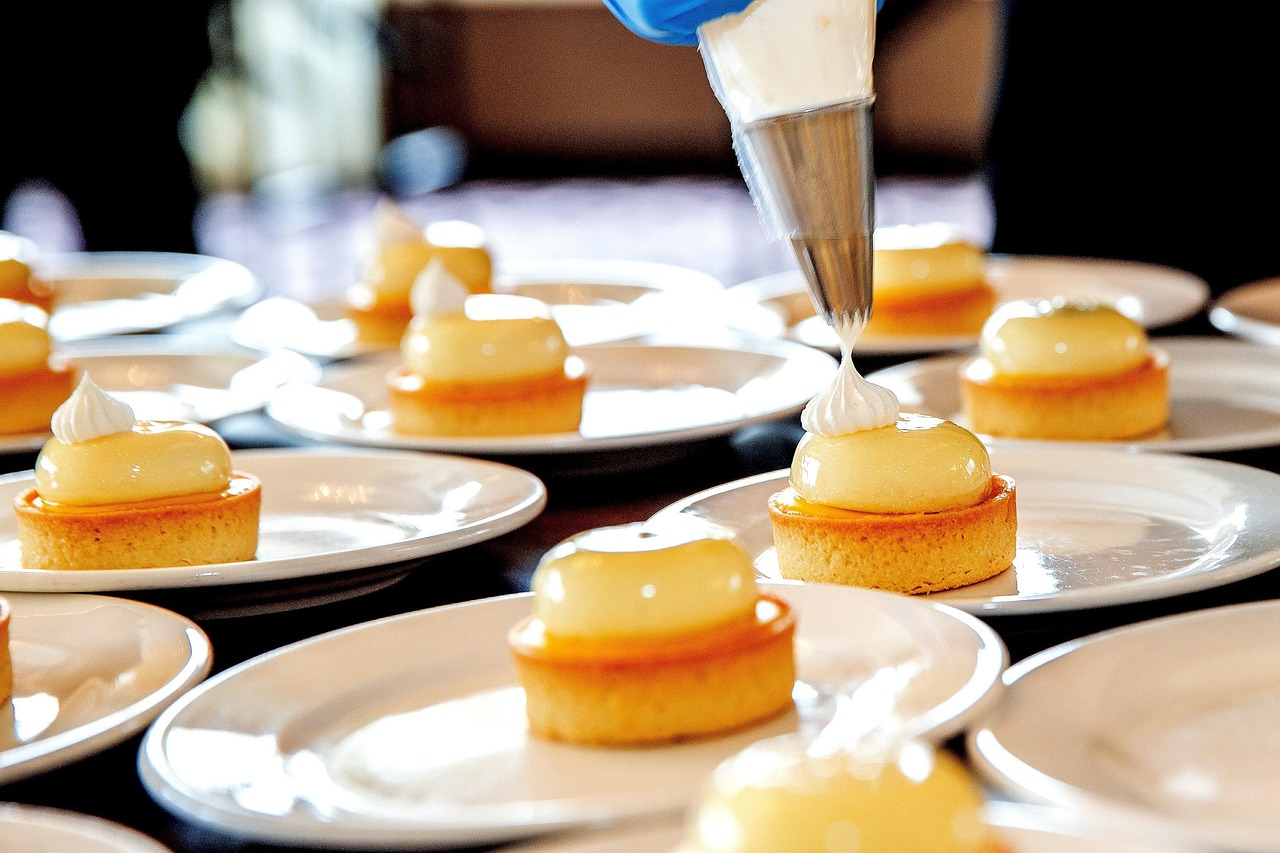 Bord met fijn gebak. Een hand met een blauwe handschoen houdt een spuitzak vast en spuit een witte massa op een taartje.