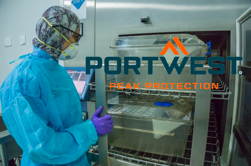 Persoon in blauwe wegwerplaboratoriumkleding voor een roestvrijstalen kast. Het logo van de fabrikant Portwest in oranje en blauw staat aan de rechterkant van de afbeelding. Een link naar onze laboratoriumkleding is bijgevoegd.