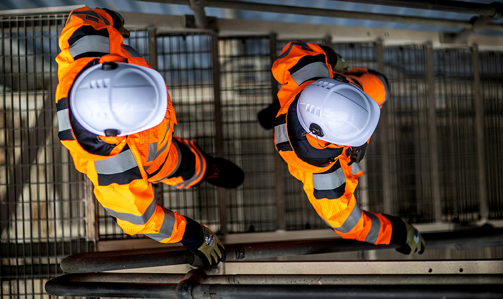 Vogelvlucht van twee arbeiders met witte helmen, zwarte en groene handschoenen, veiligheidslaarzen en oranje signalisatieoveralls met een link naar onze Arc Flash-beschermende kledingcollectie.