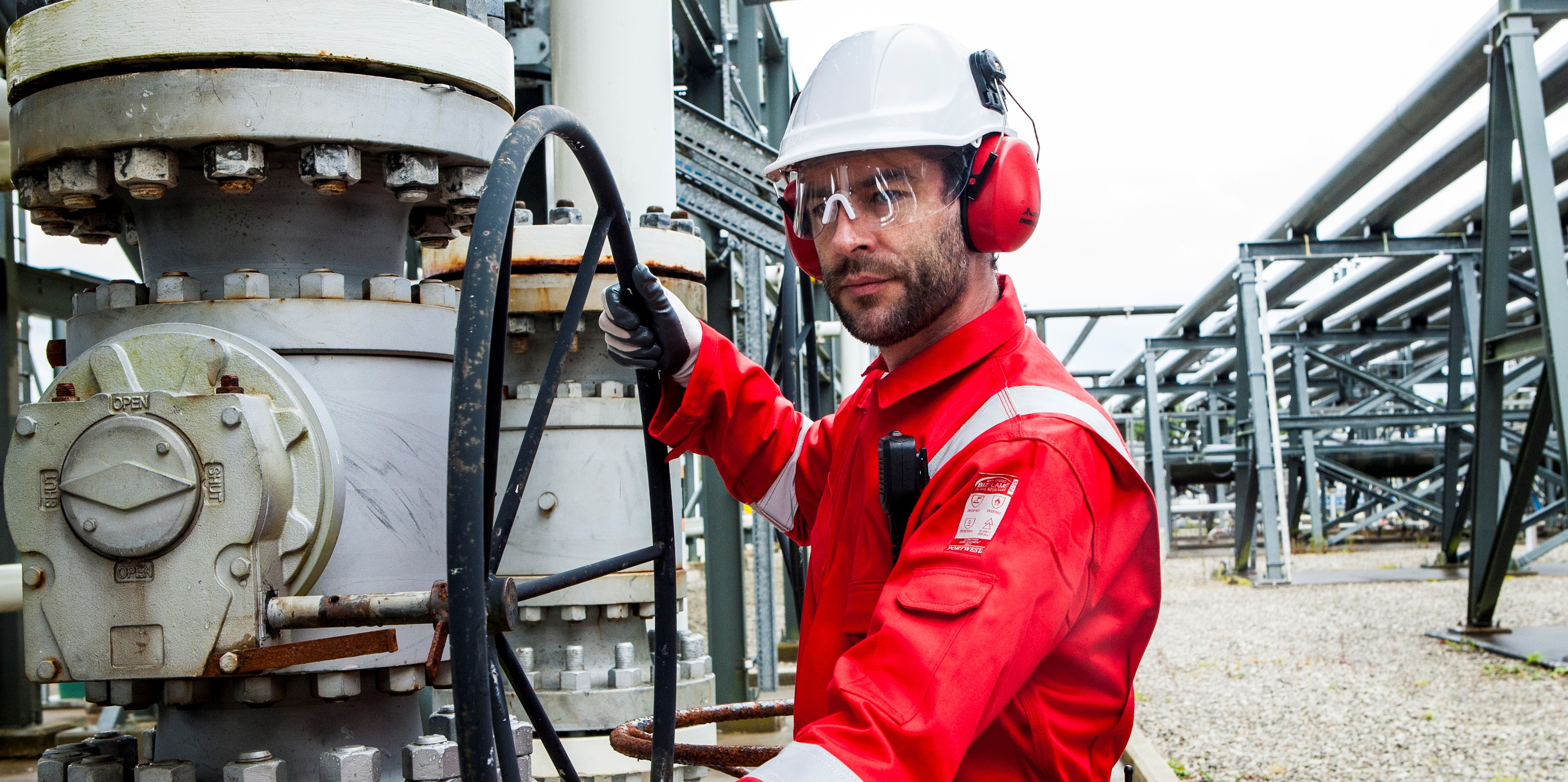 Arbeider in een industriële context met beschermende kleding: witte helm, veiligheidsbril, rode oorbeschermers, zwarte handschoenen en rode FR50-overall met reflecterende strepen.