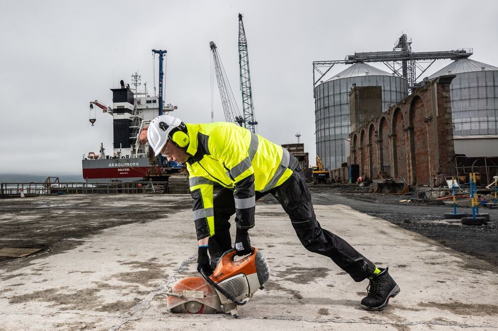 Een werknemer in een gele high-visibility jas en zwarte werkbroek die met een cirkelzaag een bodemplaat zaagt. Op de achtergrond zijn grote silo's en een containerschip te zien. Het weer is grijs en regenachtig.