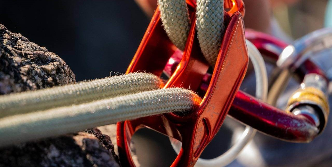 Detailopname van een oranje touwsysteem met rode en zilveren karabiners bevestigd aan een rotswand.