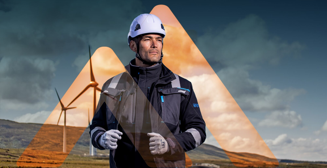 Een man met een veiligheidshelm, handschoenen en werkjas staat voor windturbines; illustratie van arbeidsveiligheid en valbeveiliging bij werken op hoogte.