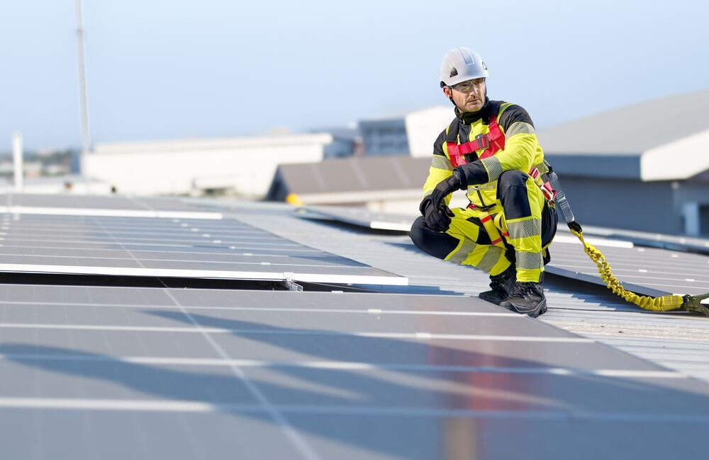 Werknemer gehurkt op een dak tussen zonnepanelen. Hij draagt fluorescerend gele werkkleding, een witte veiligheidshelm, zwarte handschoenen en schoenen. Hij is beveiligd met het Portwest 3-punts Plus FP18 harnas. Een link naar onze valbeveiliging is toegevoegd.