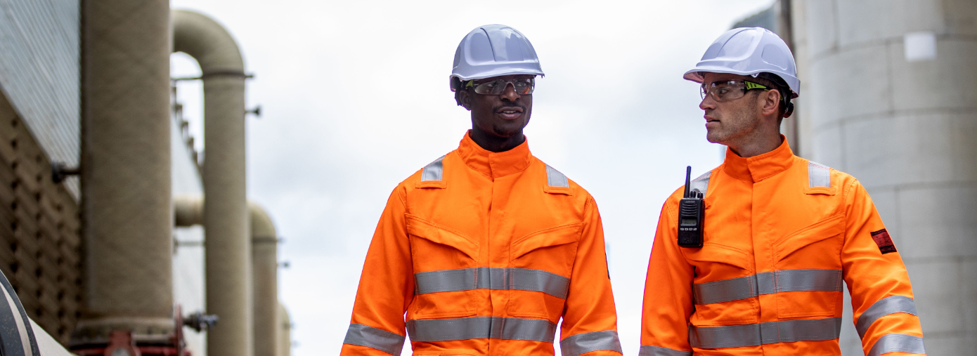 Twee werknemers voor een industriële achtergrond, gekleed in oranje hoge zichtbaarheidskleding en witte veiligheidshelmen.