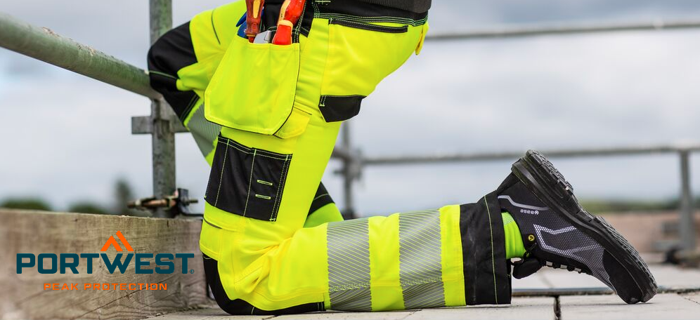 Een bouwvakker knielt op een steigervloer. Hij draagt een felgele werkbroek met hoge zichtbaarheid en geïntegreerde kniebeschermers en zwarte veiligheidsschoenen. Op de achtergrond is een bewolkte lucht te zien. Het logo van de fabrikant, Portwest, in blauwe en oranje letters staat linksonder.