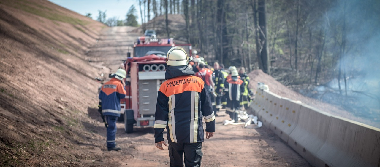 Achteraanzicht van een brandweerman in volle uitrusting. Op de achtergrond zie je een brandweerwagen op een bospad en een groep brandweermannen aan het werk. De link naar de FR98 FR98 Bizflame FR98 FR98 Bizflame FR brandvertragende overall is aanwezig.