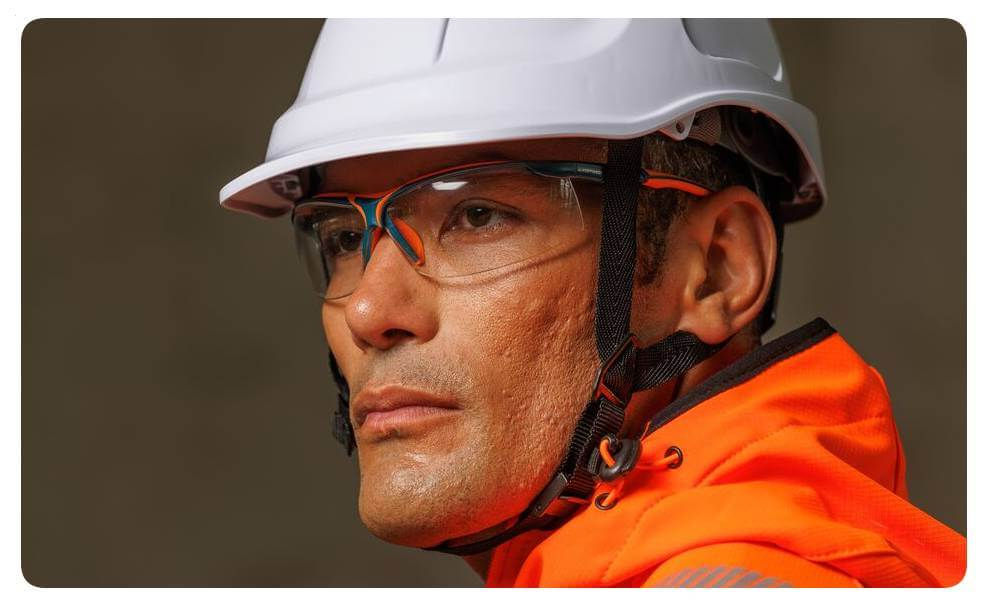 Portretopname van een man in oranje veiligheidskleding met witte veiligheidshelm en veiligheidsbril. Er is een link naar onze brillencollectie toegevoegd.