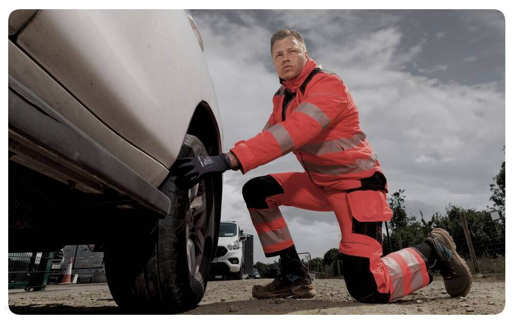 Een man in oranje high-vis werkkleding knielt voor een witte auto en werkt aan de band. Op de achtergrond is een bewolkte lucht te zien. Er is een link naar onze collectie kniebescherming toegevoegd.