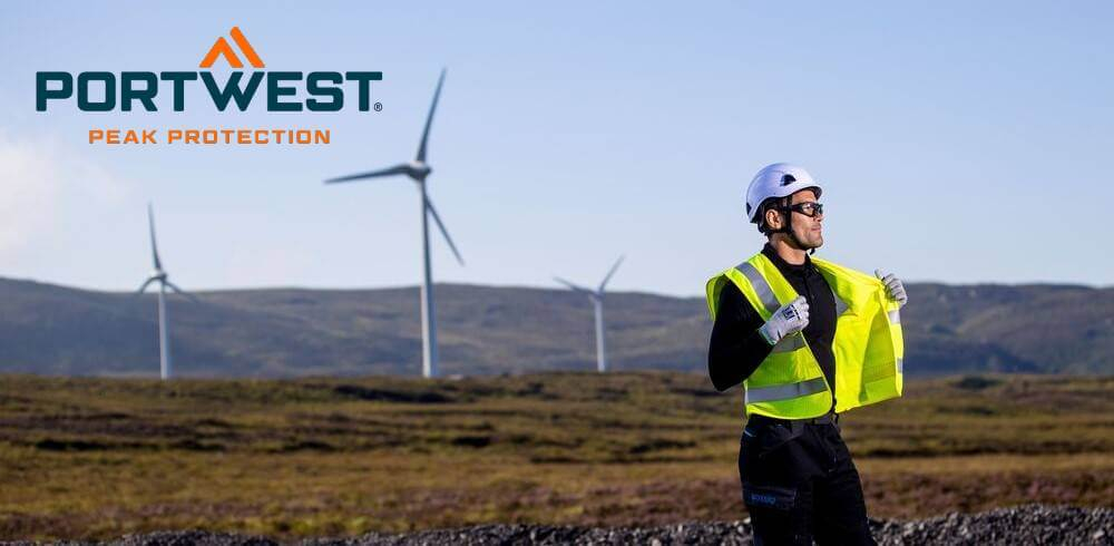 De afbeelding toont een man in zwarte werkkleding voor een kaal landschap met drie windturbines. Hij draagt een witte helm, een geel veiligheidsvest, handschoenen en een veiligheidsbril. Het Portwest-logo is in blauw en oranje zichtbaar in de linkerbovenhoek van de afbeelding.