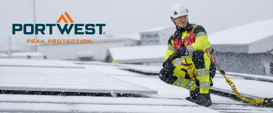 Een werknemer in een gele, goed zichtbare overall met reflecterende strepen knielt op een met sneeuw bedekt dak naast een zonnepaneleninstallatie. Hij draagt ​​een witte veiligheidshelm en valbeveiliging. Op de achtergrond zijn meer met sneeuw bedekte daken te zien en het logo van de fabrikant, Portwest, is in blauw en oranje zichtbaar linksboven in de afbeelding.