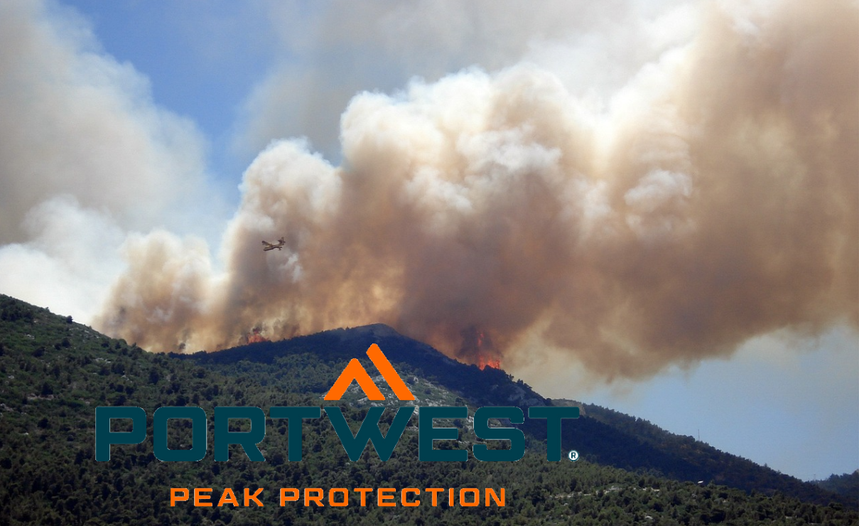 Een bosbrand woedt op een groene berghelling, dikke rookwolken stijgen op. Een blusvliegtuig nadert en werpt water of blusmiddel over de vlammen. Het logo van het merk Portwest is zichtbaar op de voorgrond – de slogan "Peak Protection" staat hieronder. Een link naar onze selectie brandweerkleding is opgenomen.