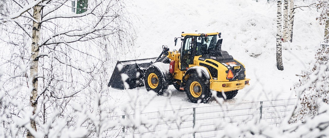 Gele tractor met schep in besneeuwd landschap met hekken en berkenbomen.
