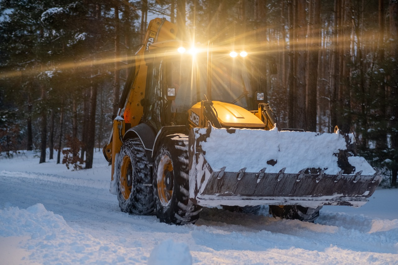 Gele trekker met een grote schep op een besneeuwde bospad. De schemering valt langzaam in en het voertuig heeft licht aan.