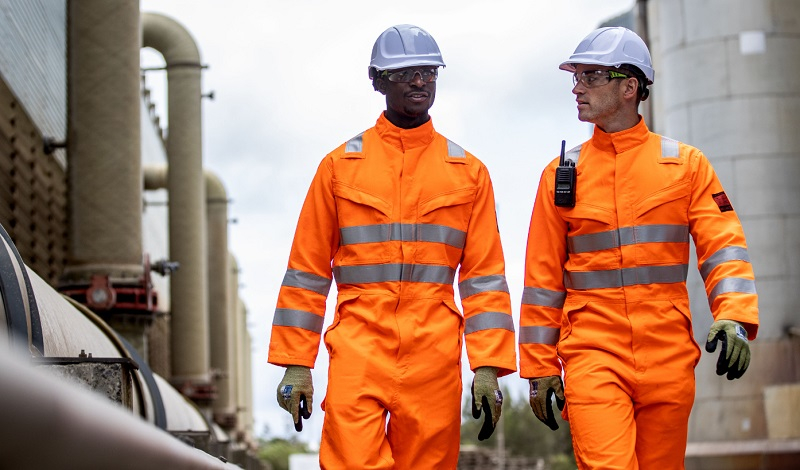 Twee werknemers voor een industriële achtergrond, gekleed in oranje hoge zichtbaarheidskleding en witte veiligheidshelmen.